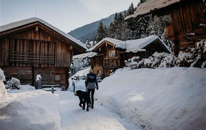 Eine Person geht mit einem Hund durch eine schneebedeckte Landschaft. Im Hintergrund sind traditionelle Holzchalets und verschneite Bäume zu sehen.