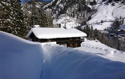 Ein malerisches Häuschen im Schnee, umgeben von hohen Tannenbäumen. Die Landschaft wird von schneebedeckten Bergen dominiert.