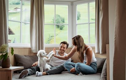 A couple is playing with a small dog in a cozy room with large windows. Outside, green hills are visible.