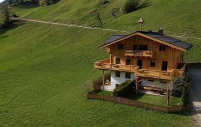 Ein schönes Holzhaus auf einer grünen Wiese. Die Umgebung ist von sanften Hügeln und einem klaren Himmel umgeben.
