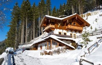 Ein gemütliches Holzhaus im Schnee, umgeben von hohen Tannenbäumen. Der Himmel ist klar und blau, und die sonnige Landschaft strahlt Ruhe aus.