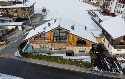 Ein modernes Haus mit Holzverkleidung und großen Fenstern, umgeben von Schnee. Im Vordergrund sieht man eine gut gepflegte Terrasse und Sträucher.
