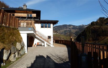 Ein modernes Haus in den Bergen mit einem gepflasterten Weg und Holzgeländer. Im Hintergrund sind grüne Hügel und ein klarer Himmel zu sehen.
