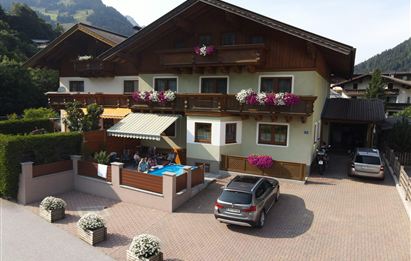 Ein gemütliches Haus mit Blumen auf den Balkonen und einer Einfahrt. Im Vordergrund sind Menschen, die sich unter einem Sonnenschirm entspannen.