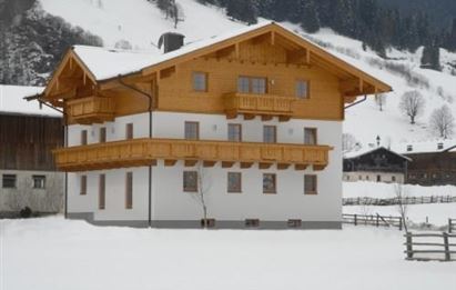 A traditional wooden house with a large balcony, surrounded by snow. The landscape is mountainous and wintry.