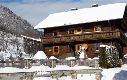 Ein schönes Holzhaus im Schnee mit einem Balkon. Die Umgebung ist winterlich, umgeben von Bergen und schneebedeckten Bäumen.