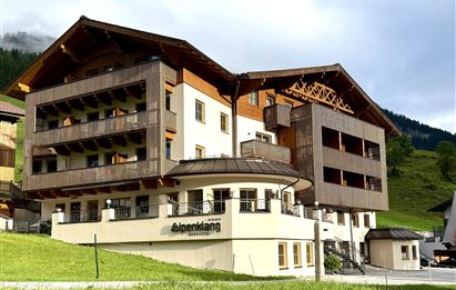 Ein modernes Hotel mit Holzfassade, umgeben von grünen Wiesen und Bergen. Der Himmel ist teilweise bewölkt, was eine ruhige Atmosphäre schafft.