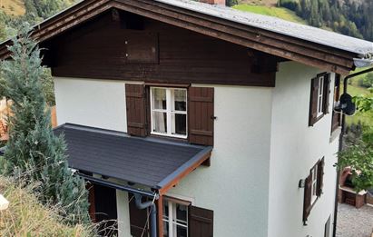 A charming two-story house in alpine style, surrounded by green mountains. The facade is light with dark wood and there is a small entrance area.