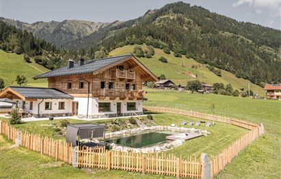 A beautiful wooden house surrounded by green meadows and mountains. In the foreground, there is a pool, surrounded by a wooden fence.