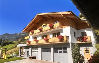 Ein schönes Ferienhaus im alpinen Stil mit blühenden Balkonkästen. Im Hintergrund sind grüne Hügel und ein blauer Himmel zu sehen.