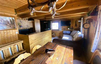 A cozy wooden apartment with a rustic dining area and a living room. In the background, a kitchen and a bright window can be seen.