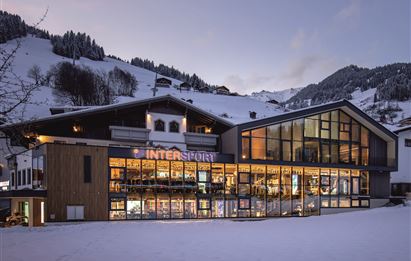 Ein modernes Sportgeschäft im Winter, umgeben von schneebedeckten Hügeln. Die große Fensterfront lässt viel Licht ins Innere und bietet einen Blick auf die Wintersportausrüstung.