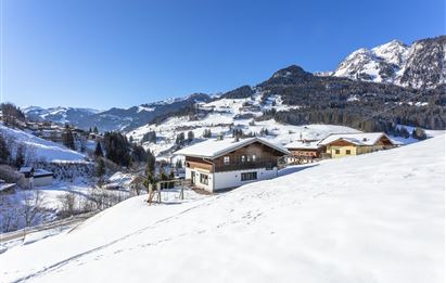 Eine verschneite Winterlandschaft mit Bergen im Hintergrund. Im Vordergrund sind einige traditionelle Holzhäuser zu sehen.