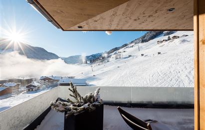 Eine herrliche winterliche Landschaft mit schneebedeckten Hügeln und einem klaren blauen Himmel. Im Vordergrund ist ein Balkon mit einem kleinen Pflanzenkübel und einem Stuhl zu sehen.