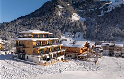 Ein modernes Gebäude im Schnee mit angrenzenden traditionellen Chalets. Im Hintergrund erhebt sich ein bewaldeter Berg unter klarem Himmel.