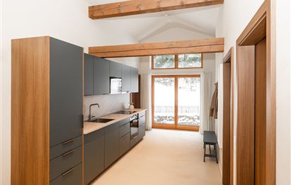 A modern kitchen with wood and gray tones. Large windows allow plenty of light in and provide a view of the structured exterior.