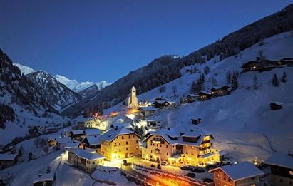 A picturesque mountain landscape with snow-covered hills and a small village that is gently illuminated. The surroundings exude a calm and cozy atmosphere.