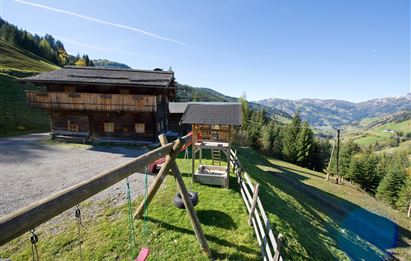 Ein schönes Holzhaus in einer bergigen Landschaft. Im Vordergrund steht ein Spielplatz mit Schaukeln und einer Rutsche.