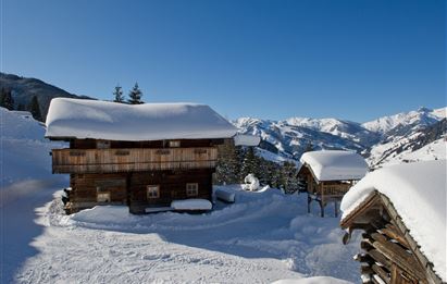 A snowy landscape with traditional wooden houses. The clear blue sky above the mountains creates a calm winter atmosphere.