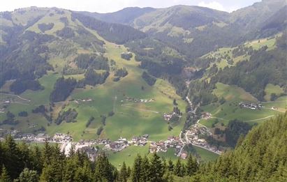 A picturesque mountain landscape with green meadows and a small village in the valley. Surrounded by tall trees and gentle hills.