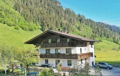 Ein malerisches Haus in den Bergen, umgeben von grünen Wiesen und Bäumen. Die Sonne scheint auf die Fassade des Gebäudes und die atemberaubende Natur.