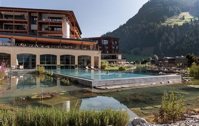 A modern hotel with an outdoor pool and lush vegetation. In the background, gentle mountains and clear skies are visible.