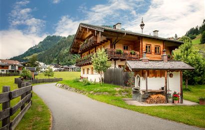 A traditional wooden house in a picturesque landscape. The path leads to the house, surrounded by green meadows and mountains.