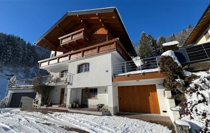 Ein modernes Haus im Schnee mit einem großen Balkon und einer Garage. Im Hintergrund sind die schneebedeckten Berge und ein blauer Himmel zu sehen.