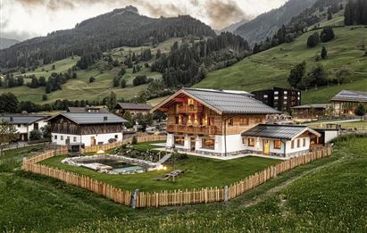 Ein schönes, alpenländisches Haus umgeben von grünen Wiesen und Bergen. Der Garten ist gut gepflegt und von einem Zaun umgeben.