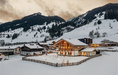 A picturesque mountain village in winter, surrounded by snow-covered hills. Cozy houses with warm light stand out against the cloudy sky.