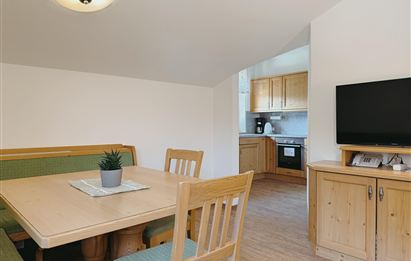 A cozy dining area with a wooden table and chairs. In the background, you can see a modern kitchenette and a television.