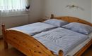 A cozy bedroom with a large wooden bed and gray bedding. The window lets in plenty of light and the walls are simple.