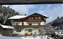 A charming house in Alpine style, surrounded by snow-covered mountains and fir trees. The sky is clear and slightly cloudy.