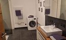 A modern bathroom with a washing machine and a large mirror. The walls are light, and there are towels in purple tones.