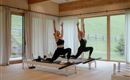 Two women are doing Pilates in a modern room with plenty of natural light. In the background, there are windows overlooking a green landscape.