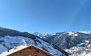 A picturesque mountain landscape with snow-covered hills and a clear blue sky. In the foreground stands a wooden house.