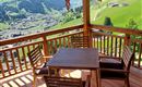 Eine Holzterrasse mit einem Tisch und Stühlen bietet einen schönen Blick auf die grüne Landschaft. Der sonnige Himmel und die Berge im Hintergrund schaffen eine einladende Atmosphäre.