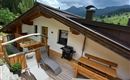 A terrace with wooden loungers and a grill, surrounded by green mountains. The sky is cloudy, perfect for relaxing hours outdoors.