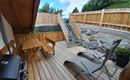 A cozy wooden terrace with a grill, a table, and lounge chairs. In the background, there is a staircase and a view of the green countryside.