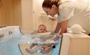 A woman is standing above a baby in a special bathtub area. The baby is safely sitting in the water and smiling, while the woman looks at it kindly.