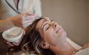 A woman is receiving a facial treatment in a relaxing environment. A hand is applying cream to her forehead.