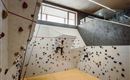 A modern climbing wall with various grips in a bright indoor space. One person is climbing on the wall, while other areas of the room are visible.
