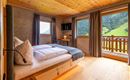 Ein gemütliches Schlafzimmer mit Holzdecke und großen Fenstern. Der Blick zeigt eine grüne Landschaft mit Bergen und Blick auf den Balkon.