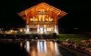 Ein wunderschönes Holzhaus bei Nacht, beleuchtet von warmem Licht. Im Vordergrund spiegelt sich das Haus im ruhigen Wasser eines kleinen Teichs.
