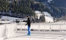 A man stands on an ice surface in a wintry landscape. Snow-covered mountains and forests can be seen in the background.