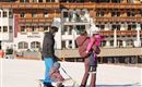 A family stands in the snow while pulling a sled. Beautiful mountain houses are visible in the background.
