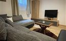 Ein gemütliches Wohnzimmer mit einem grauen Sofa und einem Holztisch. Ein Fernseher steht auf einem Möbelstück im Hintergrund.