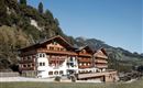 A cozy hotel in Alpine style, surrounded by green meadows and mountains. The sky is clear and bright blue.
