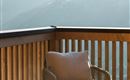 Ein bequemer Sessel auf einem Balkon mit Holzgeländer. Im Hintergrund sind sanfte Hügel und Berge vor einem hellen Himmel zu sehen.