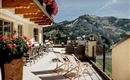 Eine sonnige Terrasse mit gemütlichen Liegestühlen und blühenden Blumen. Im Hintergrund sind die beeindruckenden Berge sichtbar.